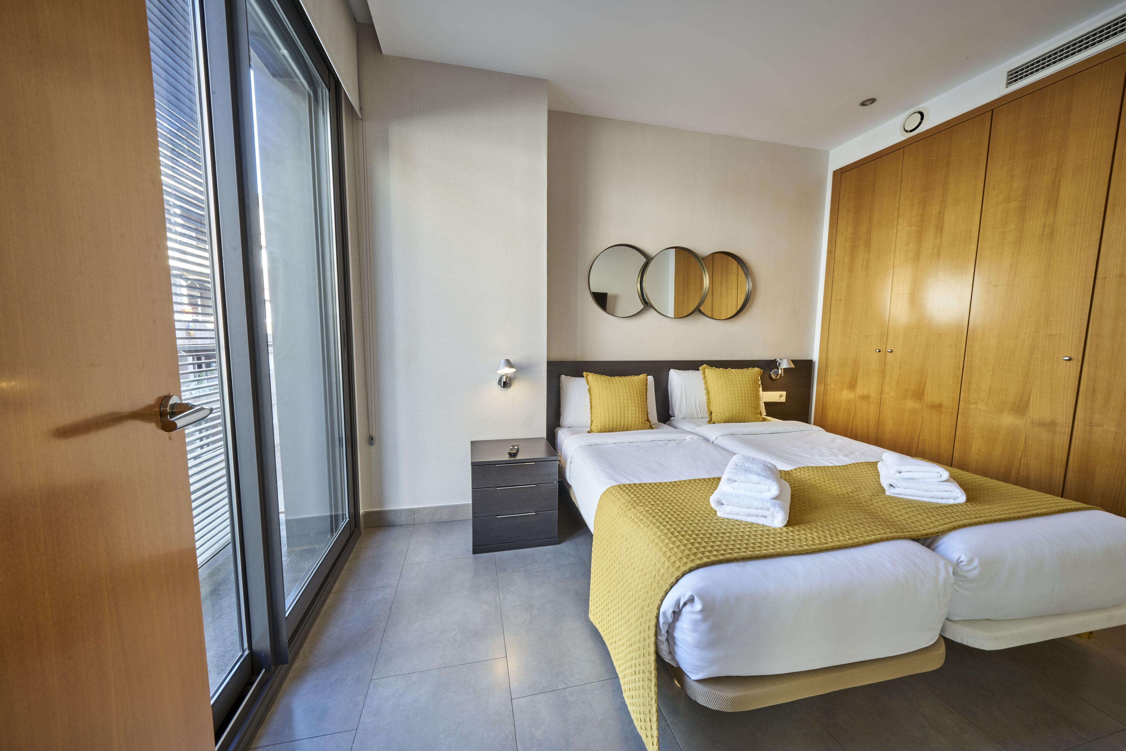 Una habitación de hotel moderna con una cama grande, una mesita de noche, un armario y un ventanal amplio.