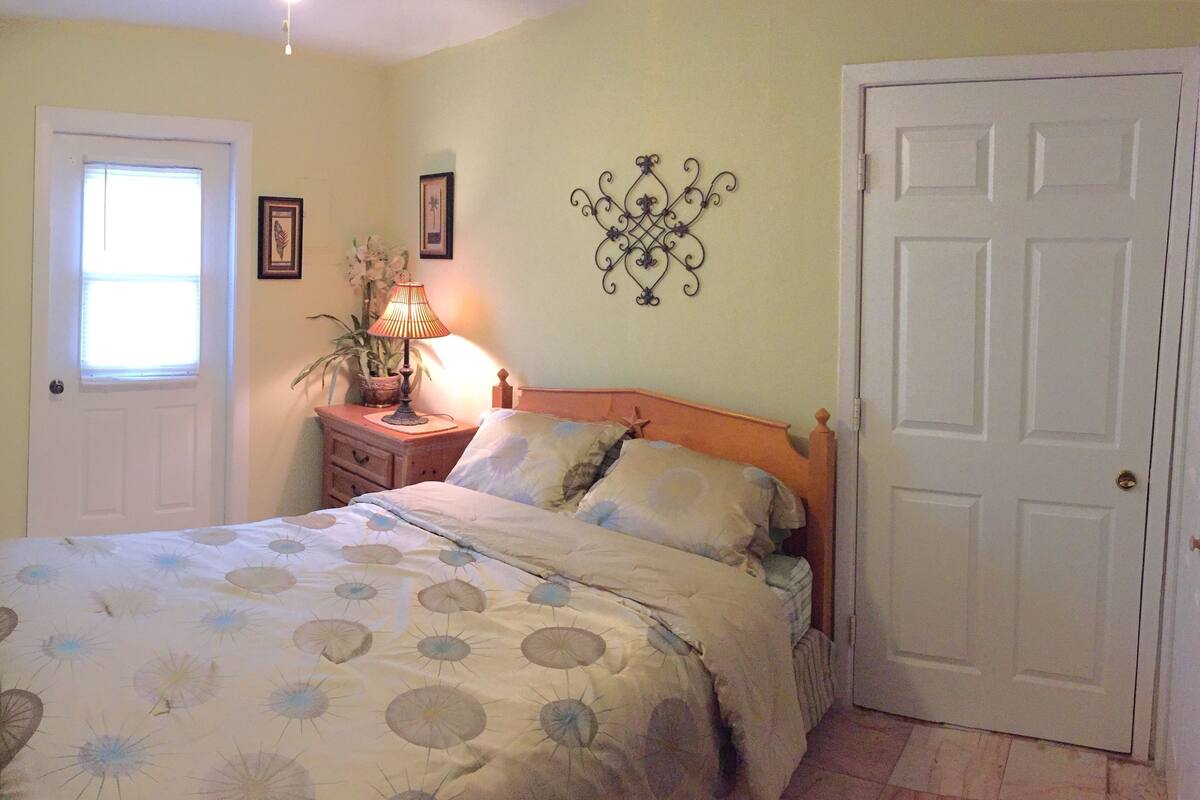 A bedroom with a bed, a nightstand with a lamp, a potted plant, and a door.