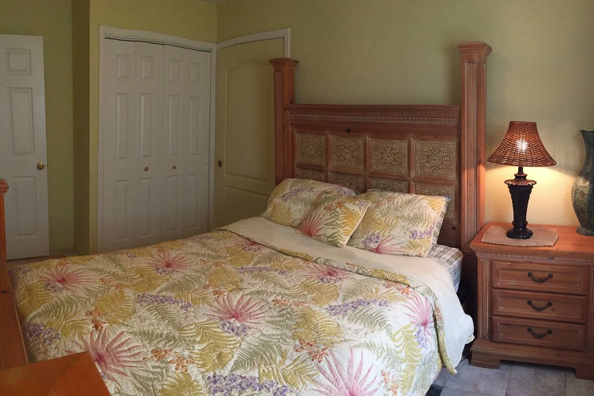 A bedroom with a floral bedspread, a wooden headboard, a bedside lamp, a nightstand with drawers, and two doors.
