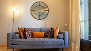 Un sofá gris con cojines de colores, un espejo redondo y una lámpara de pie en una habitación con ventana y cortinas.