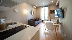 Una cocina moderna con estufa integrada, un comedor con mesa y sillas, un sofá gris con espejo y un televisor montado en la pared.