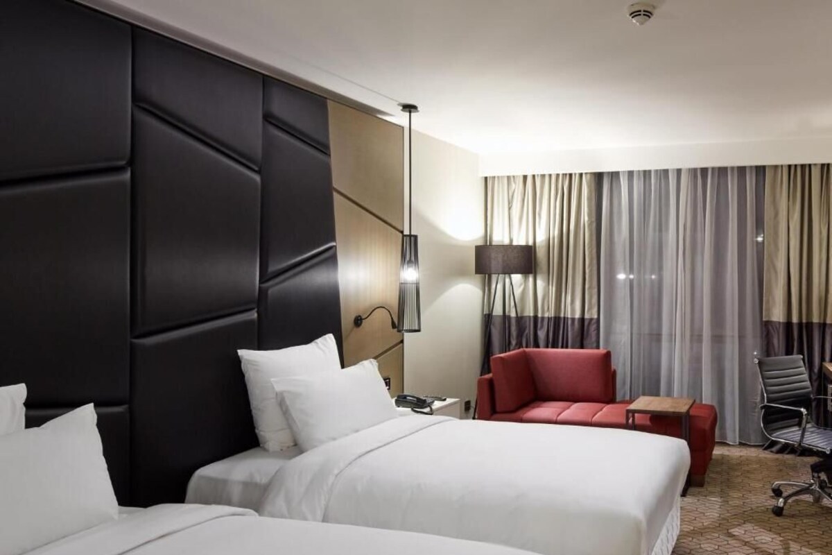 Une chambre d’hôtel avec deux lits, un fauteuil rouge, un bureau et une grande fenêtre avec des rideaux.