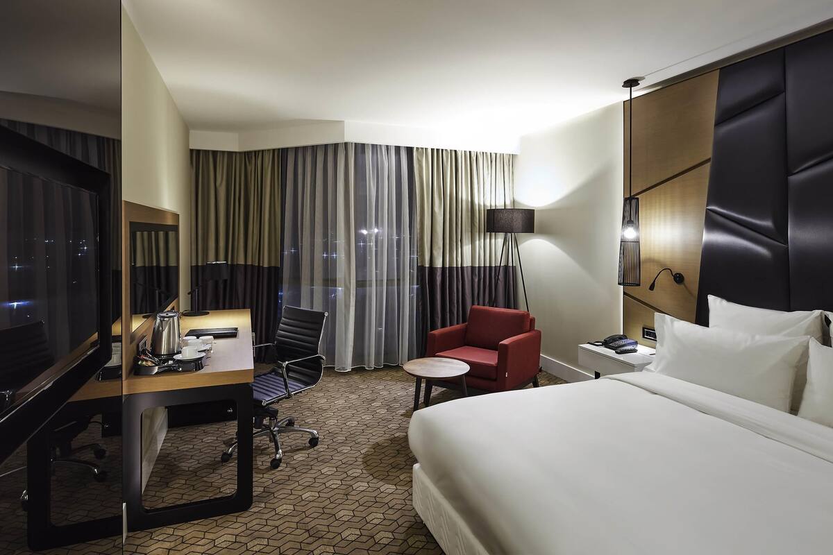 Een hotelkamer met een bed, een bureau met stoel, een televisie, een lamp en een rode fauteuil.