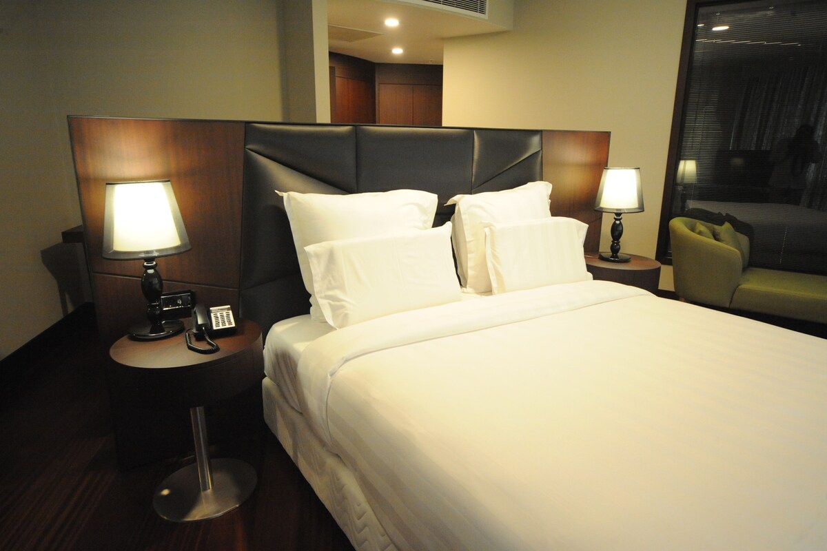 Une chambre d’hôtel avec un grand lit, deux tables de chevet avec des lampes, un téléphone et un fauteuil vert.