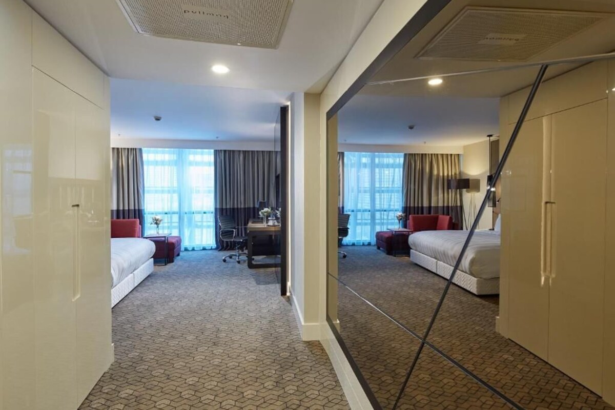 Une chambre d’hôtel avec un grand lit, un bureau et une chaise. On y trouve une fenêtre avec des rideaux, un miroir et une armoire.