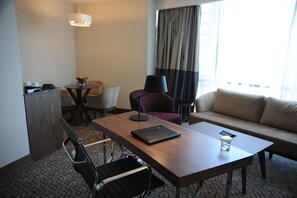 Une chambre d’hôtel avec une table à manger en bois, un canapé, une chaise, une petite table avec une lampe et une fenêtre avec des rideaux.