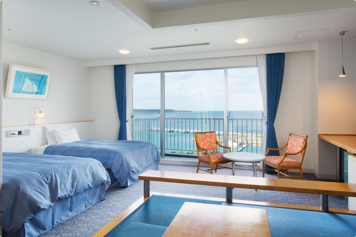 A hotel room with two beds, a balcony with a view of the sea, a small table, and a chair.