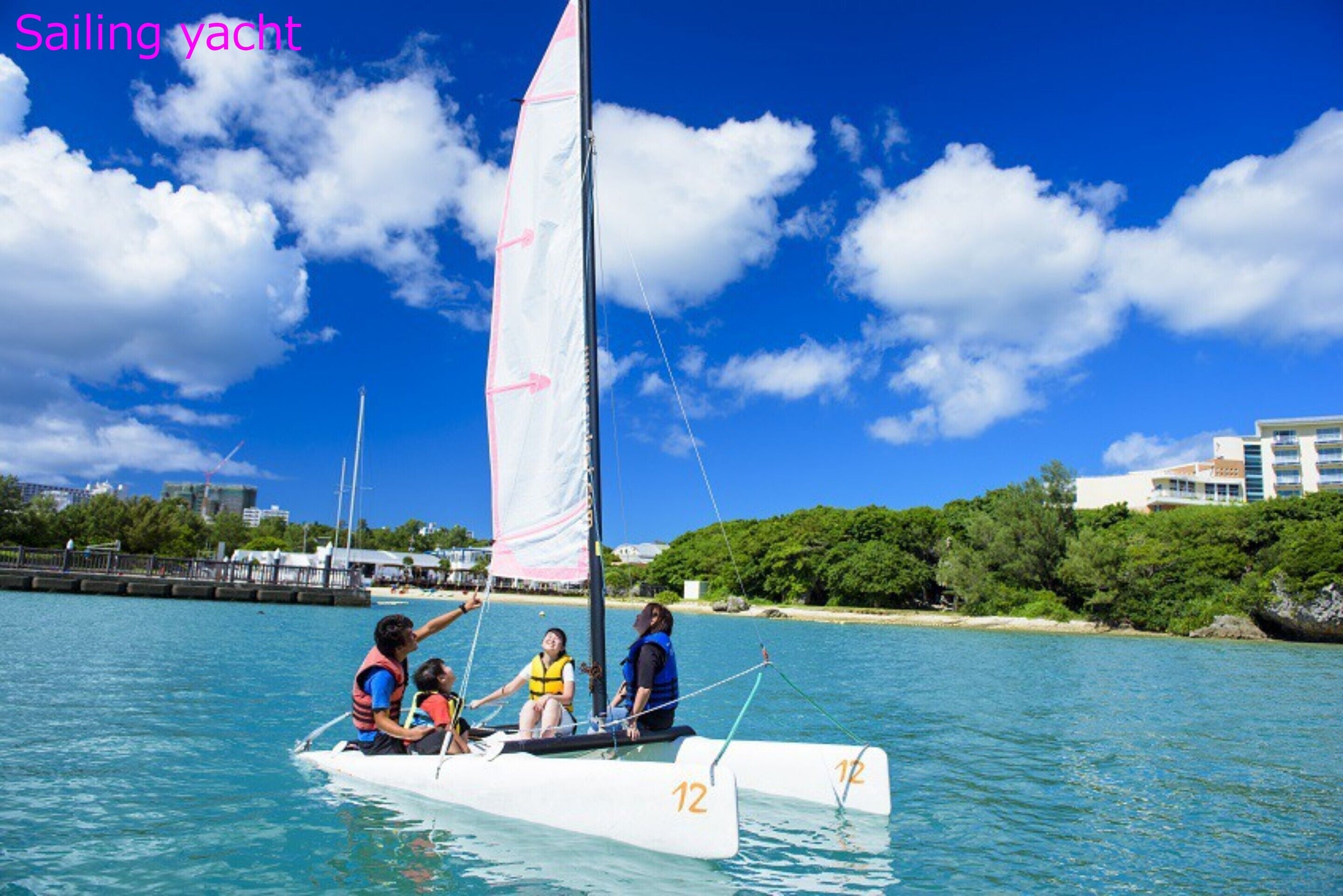 A sailing yacht with four people on board, a clear blue sky, and a coastal landscape with buildings in the background.