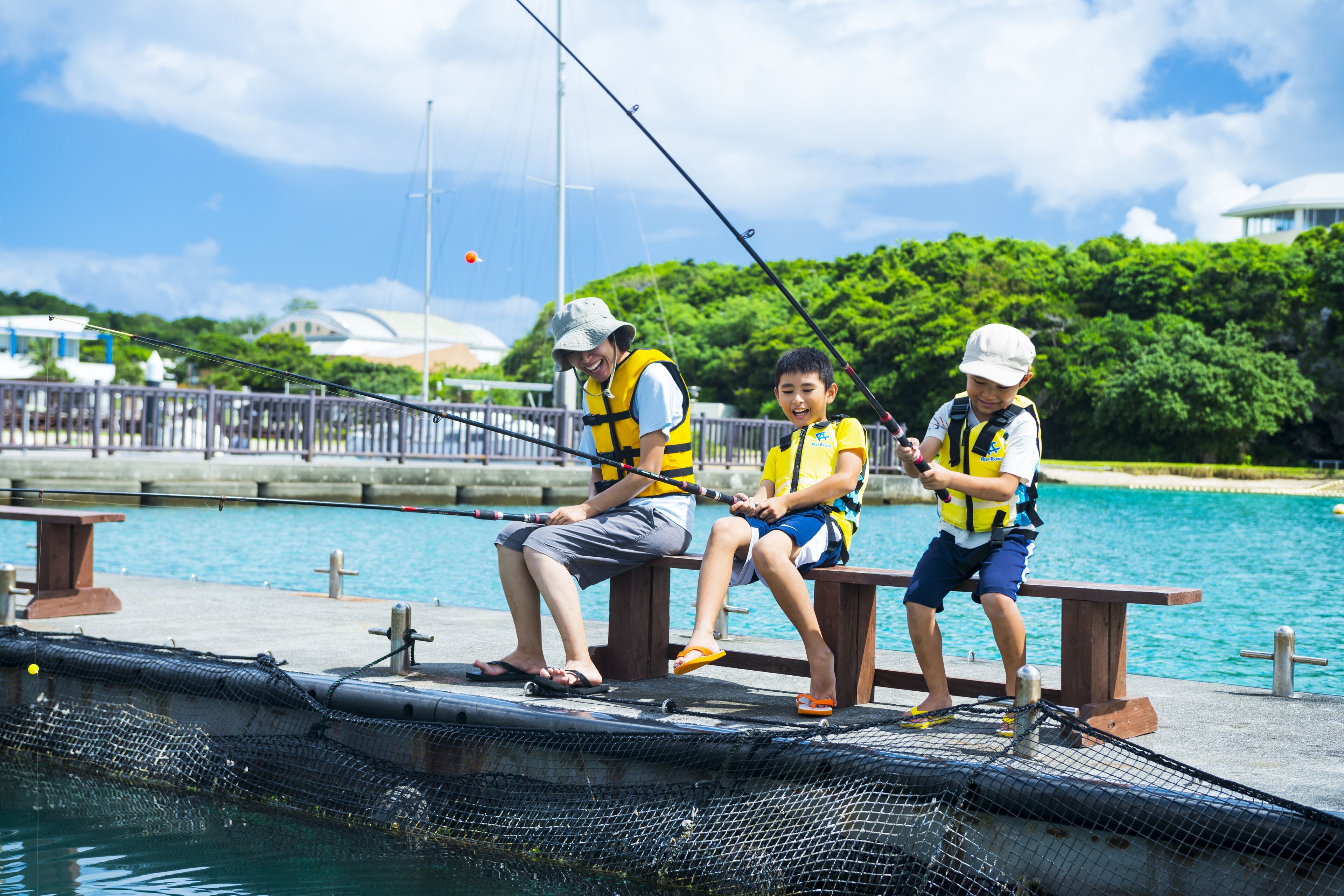 Three people sitting on a dock by a body of water, wearing life jackets and fishing rods.