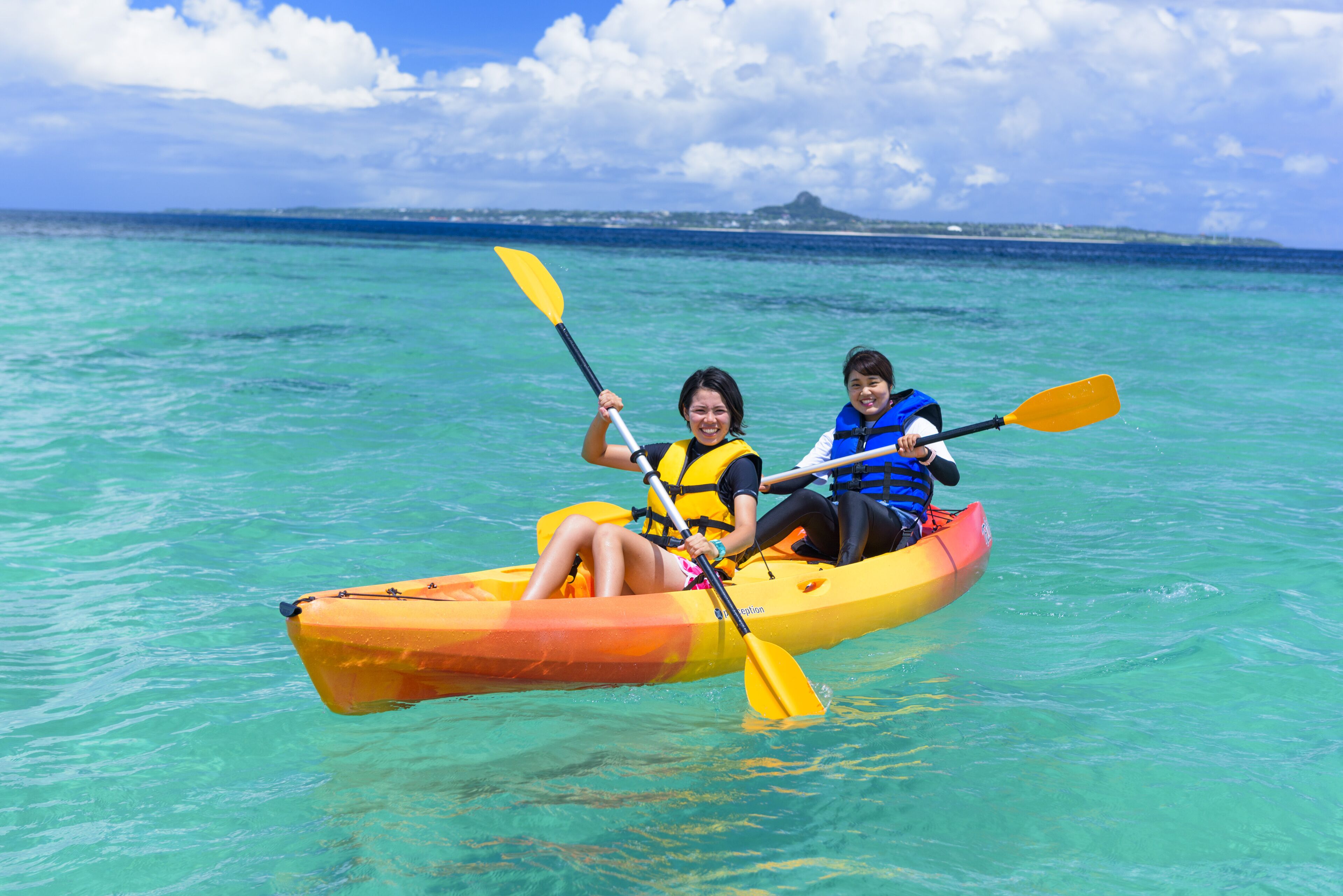 Two people kayaking on clear turquoise water with a distant island and a mountain in the background.