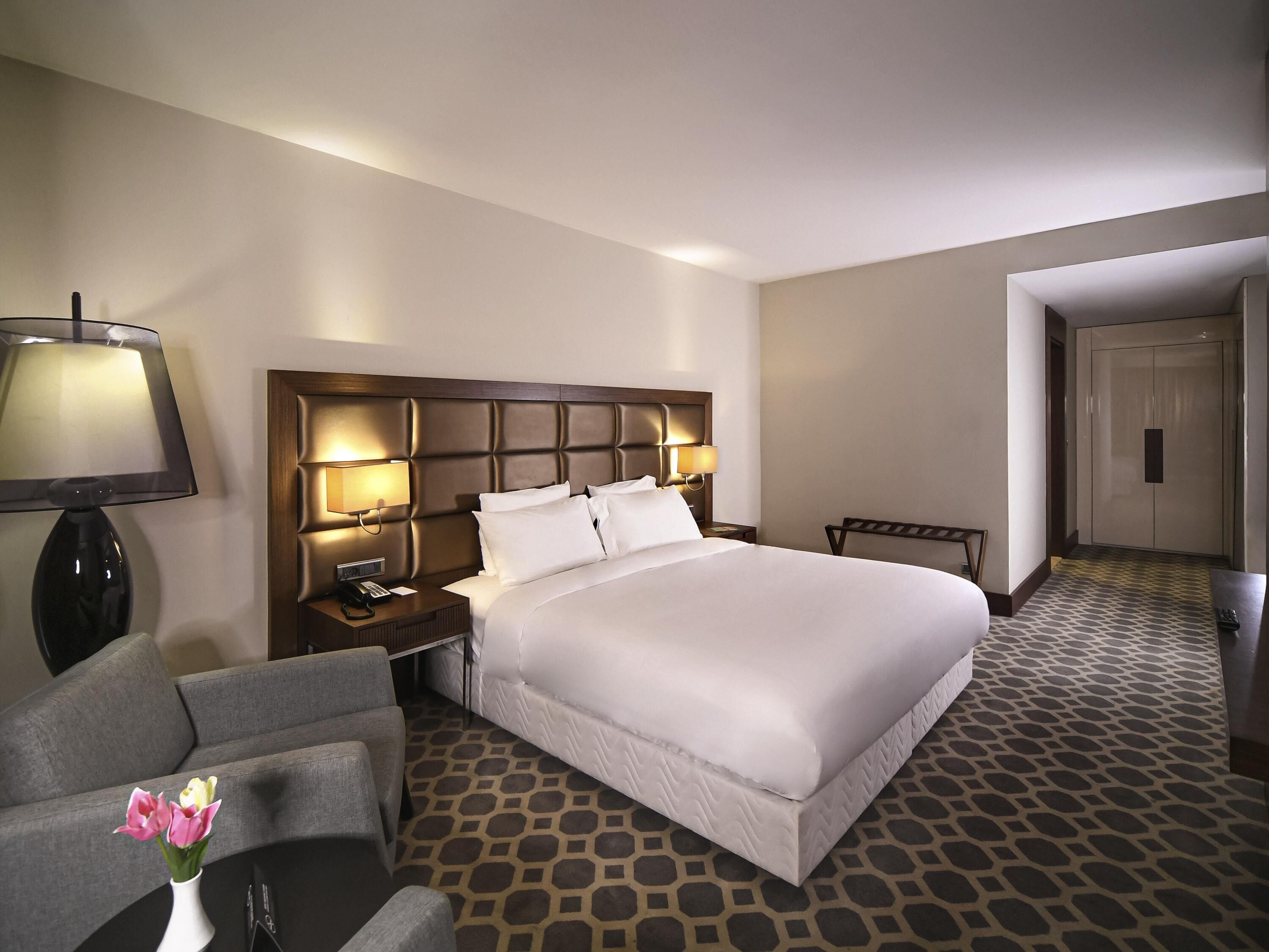 A hotel room with a large bed, a bedside table with a lamp, a chair, and a vase with flowers.