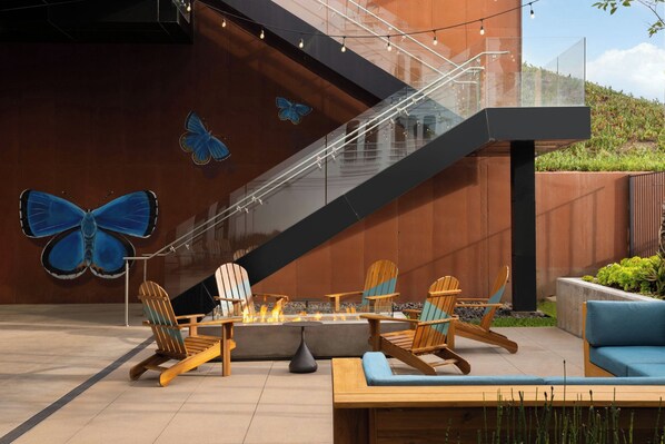 A modern outdoor seating area with Adirondack chairs, a fire pit, and a staircase against a wall with butterfly murals. - Aloft by Marriott El Segundo - Los Angeles Airport (El Segundo)