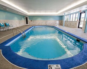 An indoor swimming pool with a 5 ft depth limit sign, blue poolside chairs, and a clear view of the outside through large windows.