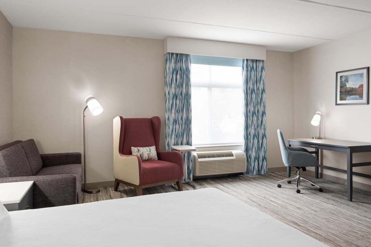 A hotel room with a bed, a desk, a chair, a red armchair, a floor lamp, and an air conditioning unit.