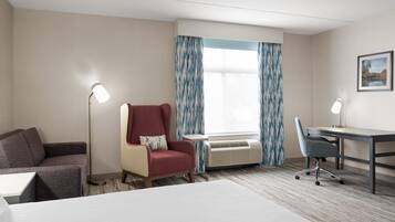 A hotel room with a bed, a desk, a chair, a red armchair, a floor lamp, and an air conditioning unit.