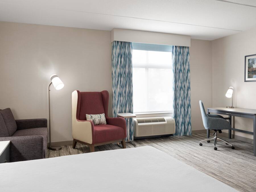 A hotel room with a bed, a desk, a chair, a red armchair, a floor lamp, and an air conditioning unit.