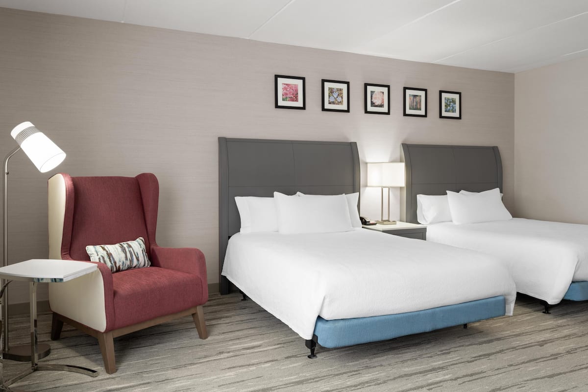 A hotel room with two beds, a red armchair, a small table, and a floor lamp.