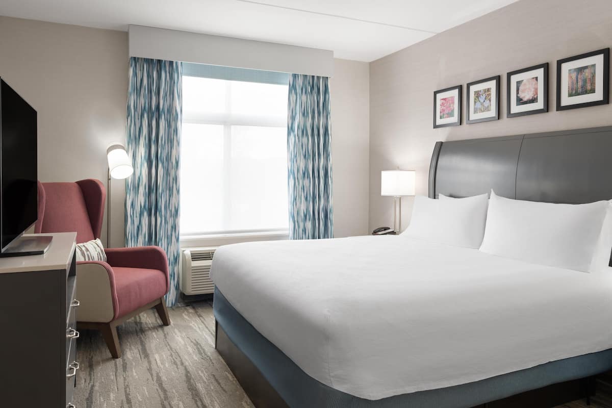 A hotel room with a bed, a desk, a chair, a lamp, and a window with curtains.