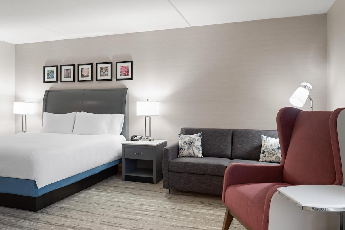 A hotel room with a bed, a sofa, a chair, a nightstand, and a lamp.