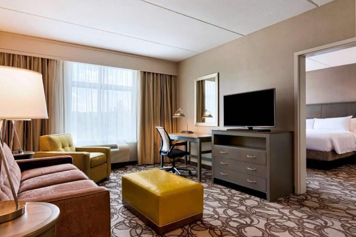 A hotel room with a sofa, a desk with a TV, a chair, a bed, and a large window.