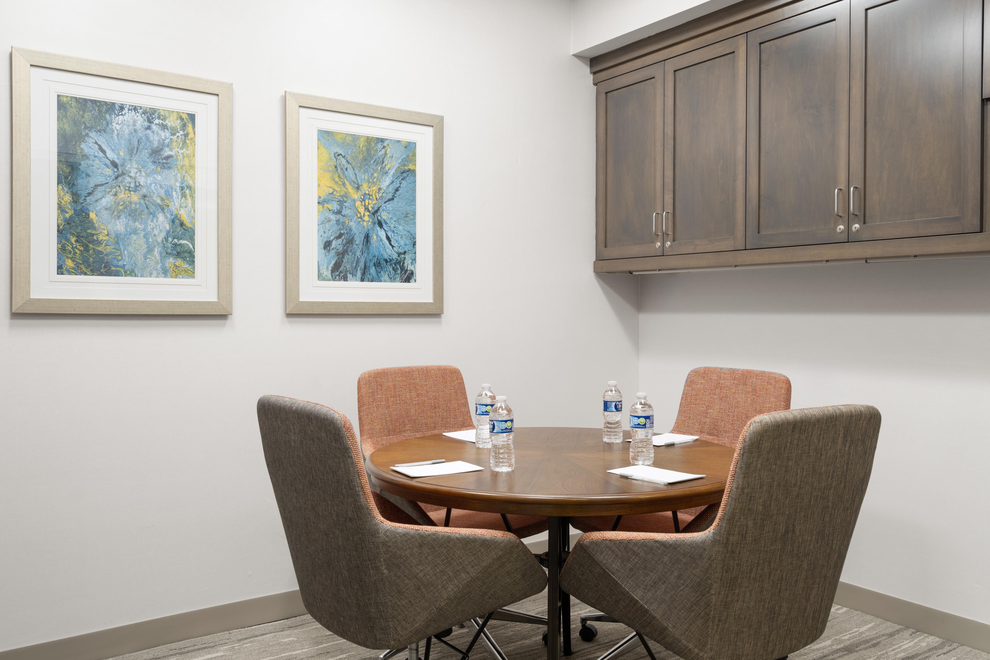 A conference room with a round table, four chairs, two framed abstract paintings, and a wooden cabinet.