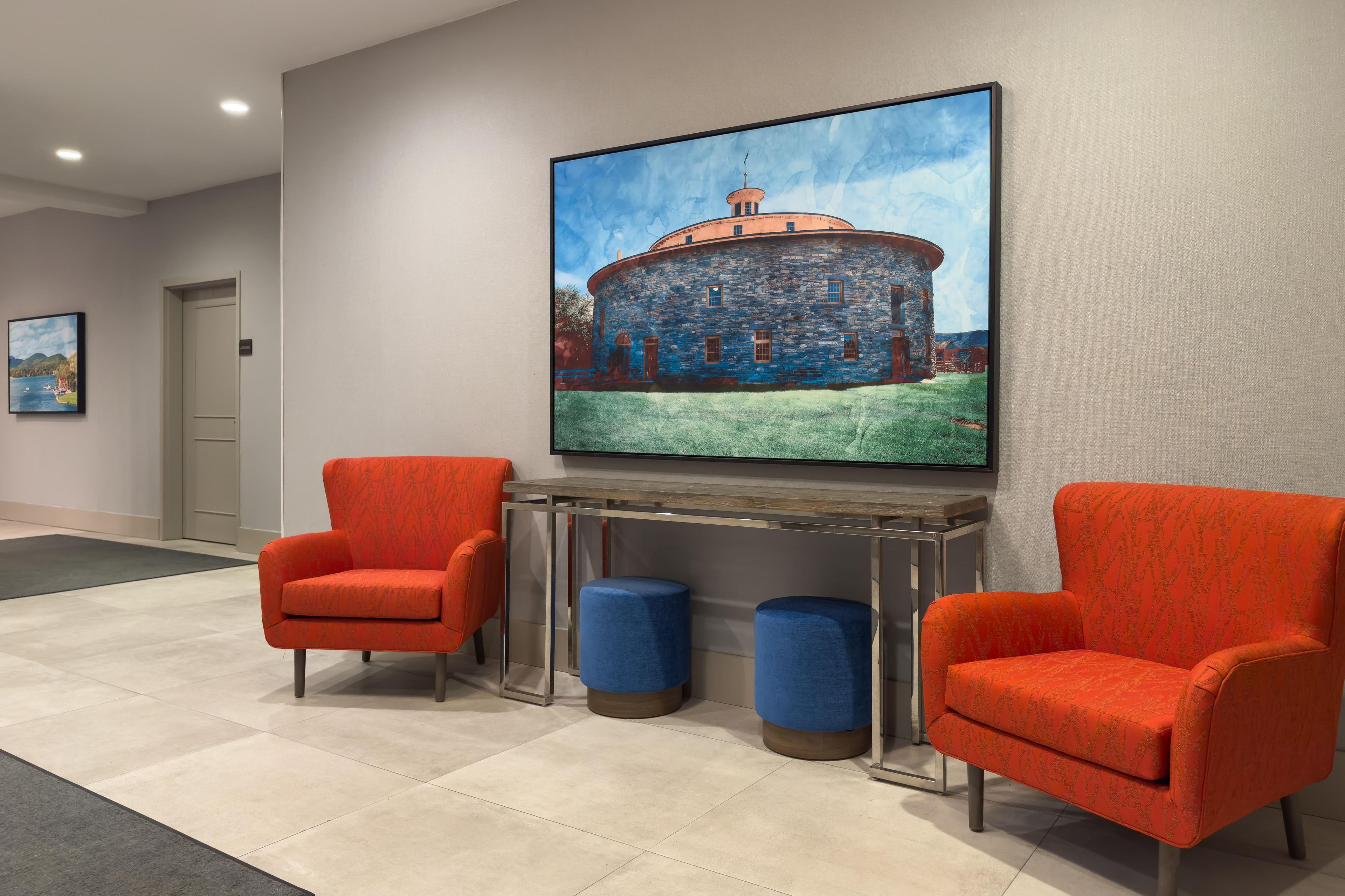 A lobby with two orange armchairs, a large painting of a stone building, and a small table with blue stools.