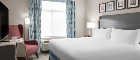 A hotel room with a bed, a desk with a laptop, a chair, a bedside table with a lamp, and a window with curtains.