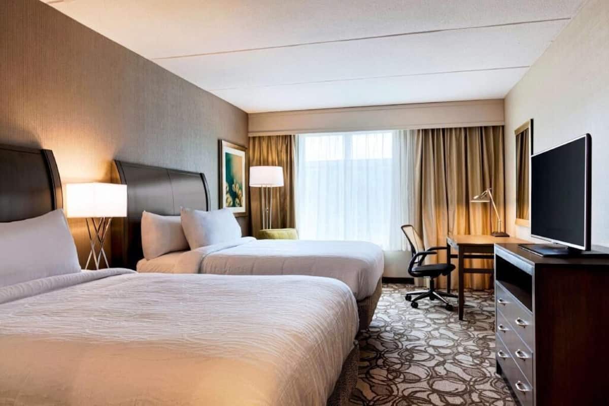 A hotel room with two beds, a desk, a television, and a window with curtains.