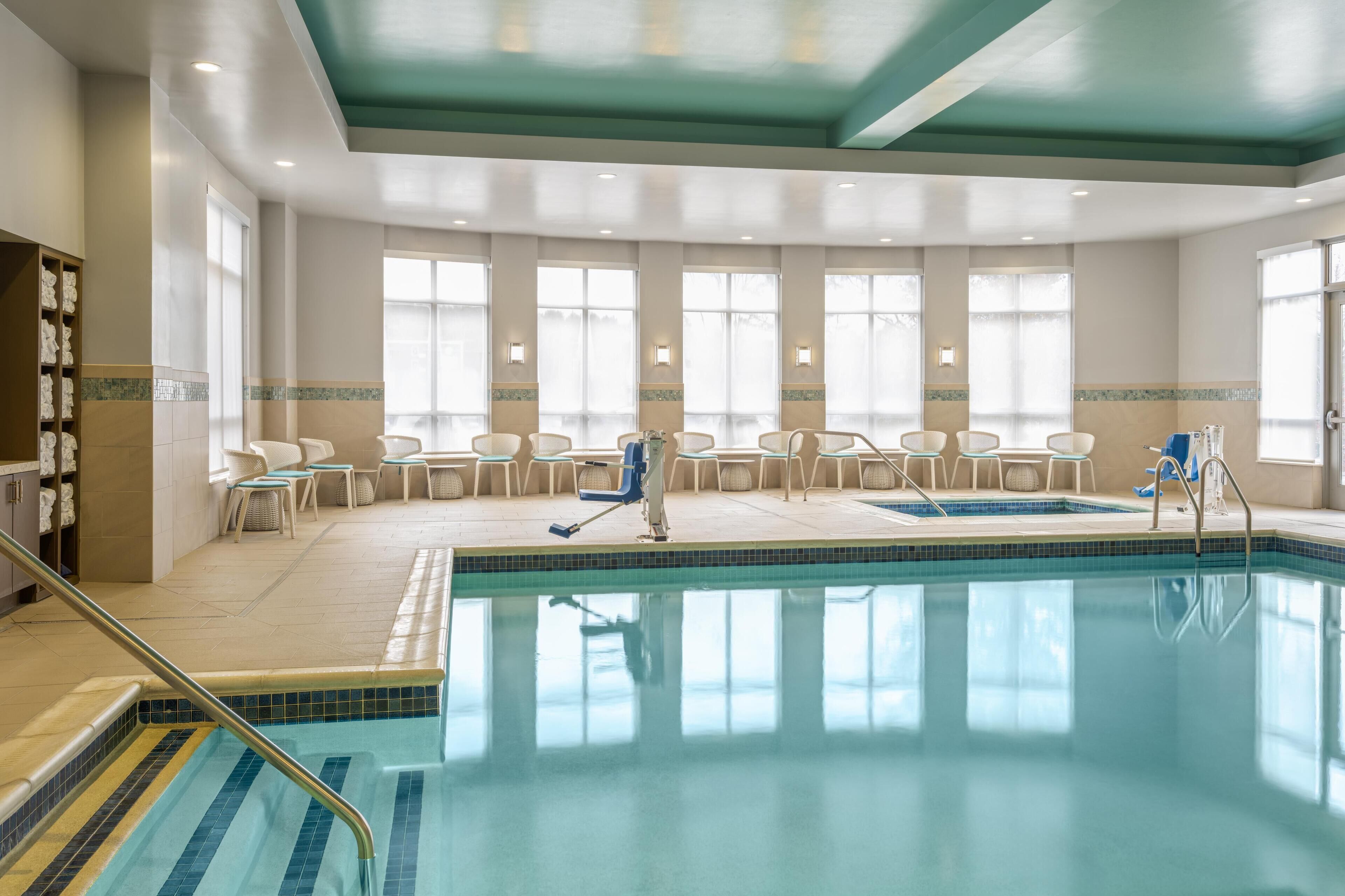 Une piscine intérieure à l’eau d’un bleu limpide, entourée de chaises longues et de tables, et dotée de grandes fenêtres qui laissent entrer la lumière naturelle.