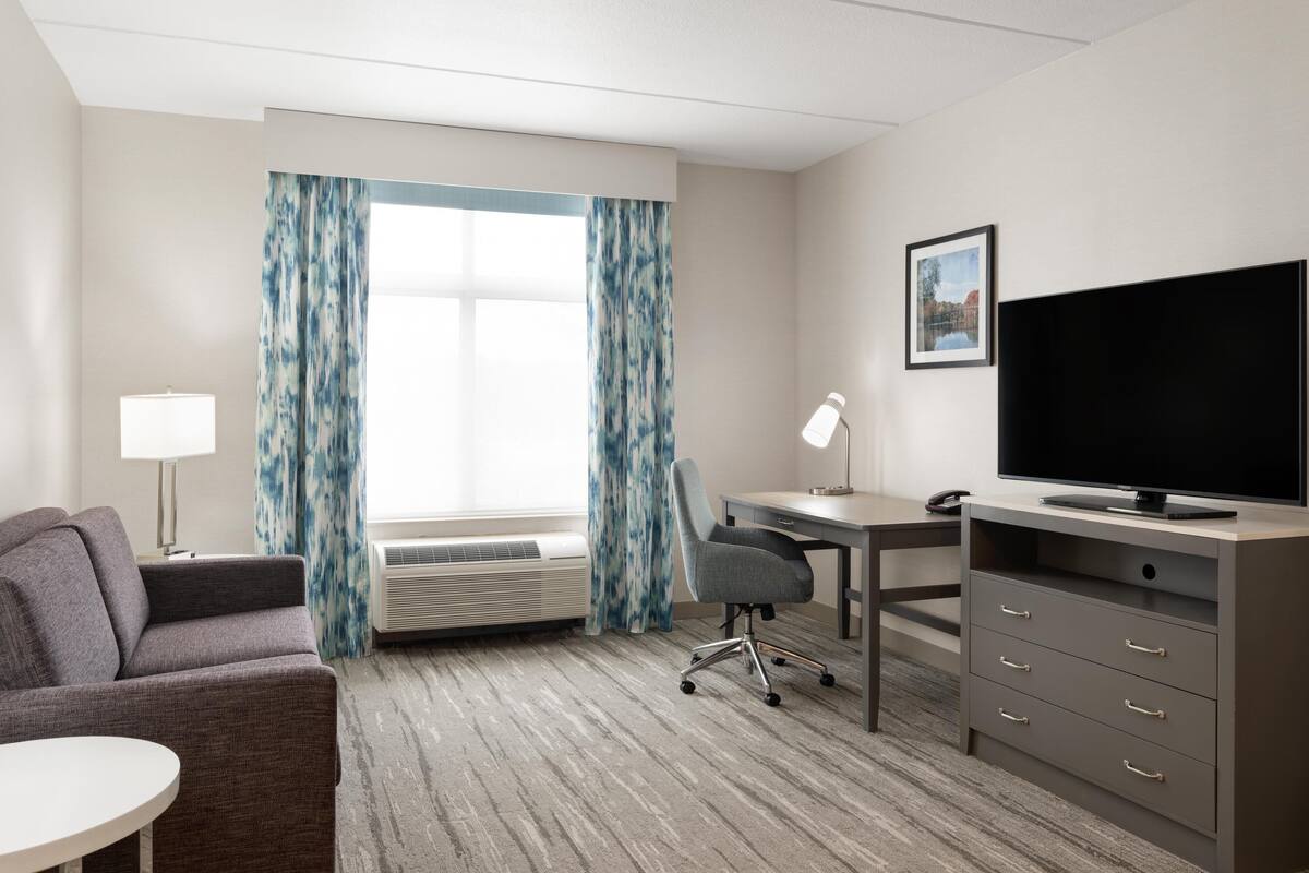 A hotel room with a gray sofa, a desk with a computer, a flat-screen TV, a lamp, and a window with blue curtains.
