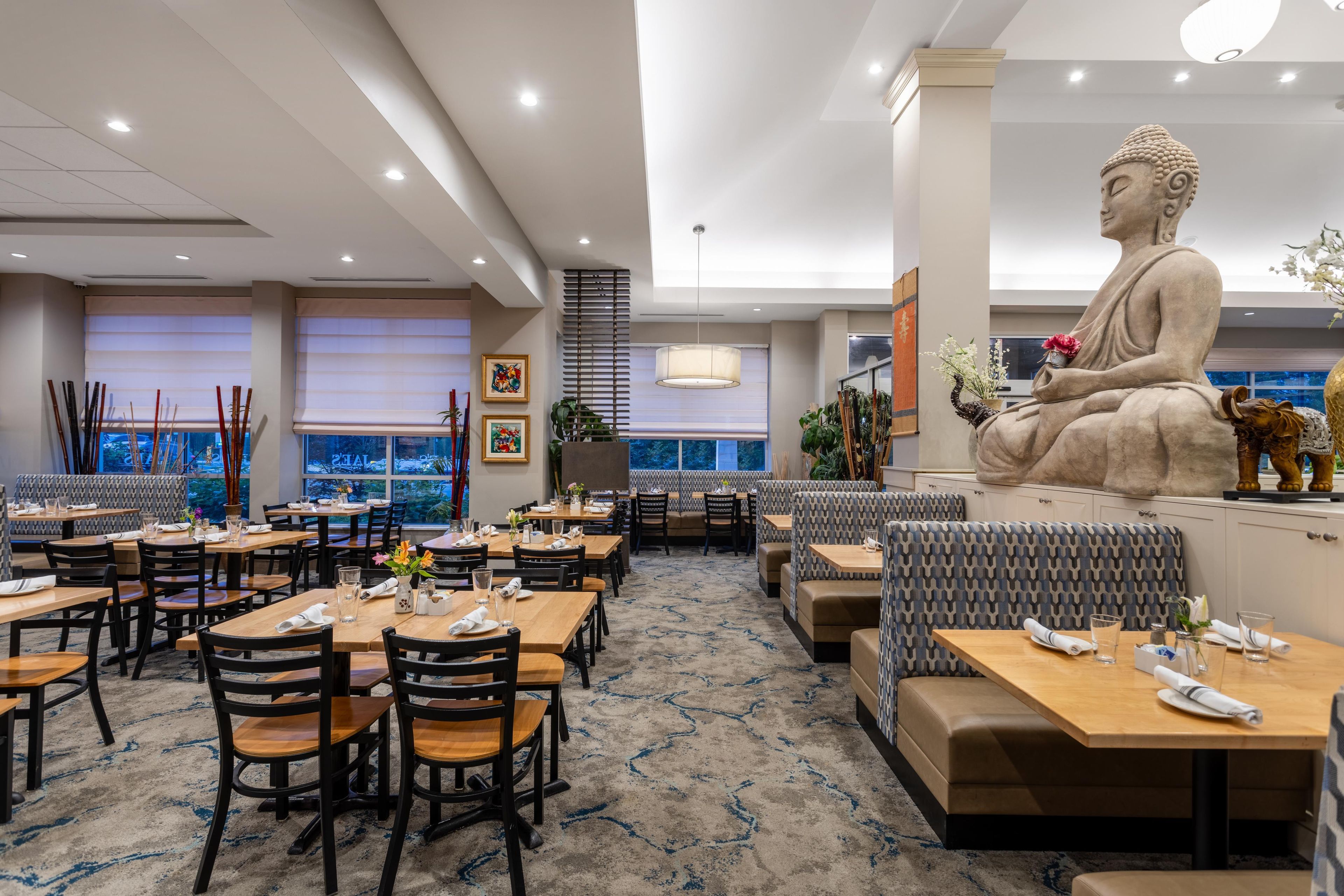 Un restaurant doté de nombreuses tables et de plusieurs coins repas, d’une grande statue de Bouddha et offrant une vue sur le paysage urbain grâce à de grandes fenêtres.