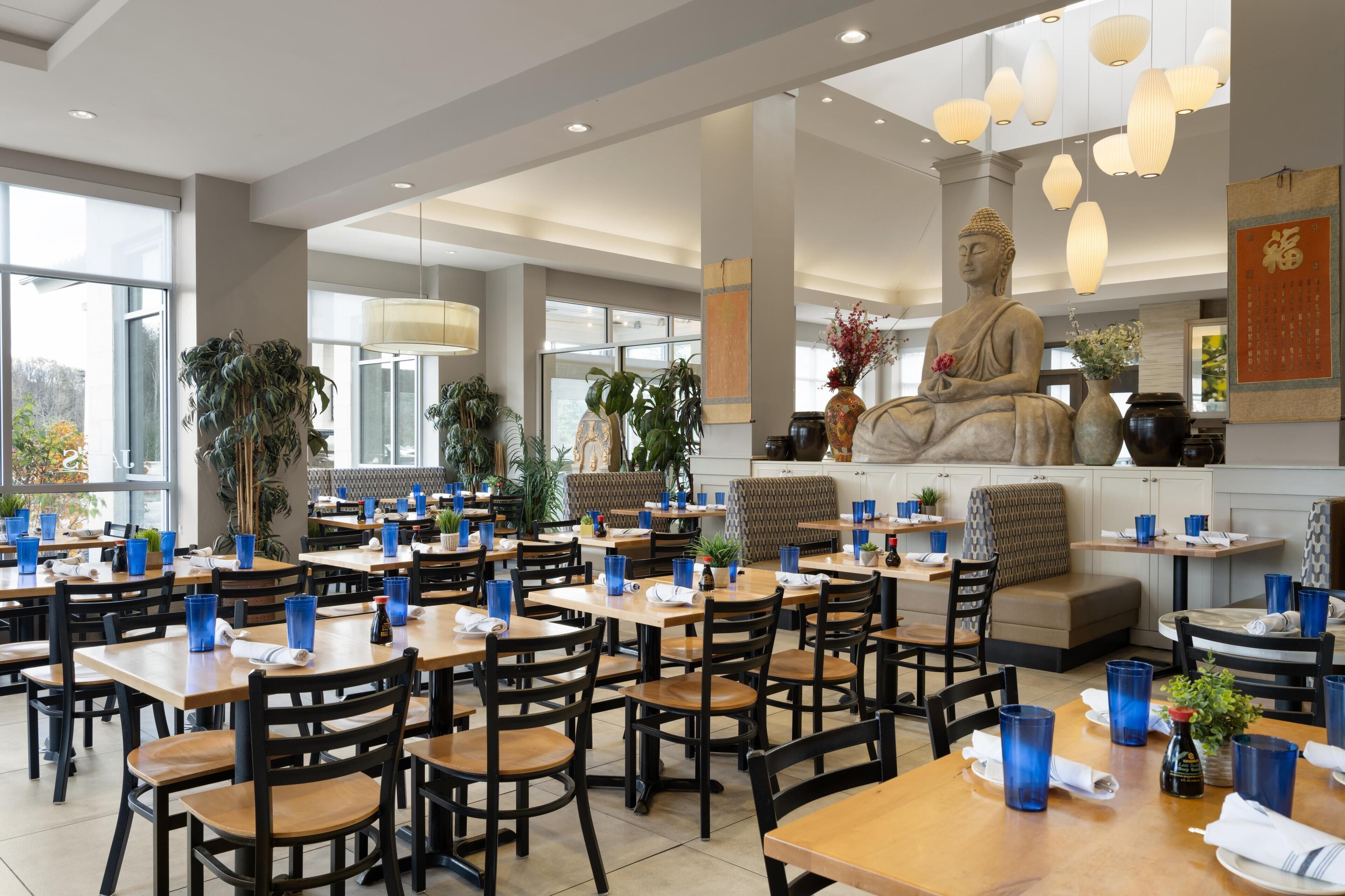 A restaurant with multiple tables and chairs, a Buddha statue, and various plants.