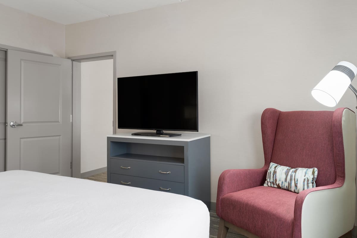 A hotel room with a bed, a television on a stand, a desk with drawers, a chair with a pillow, and a lamp.