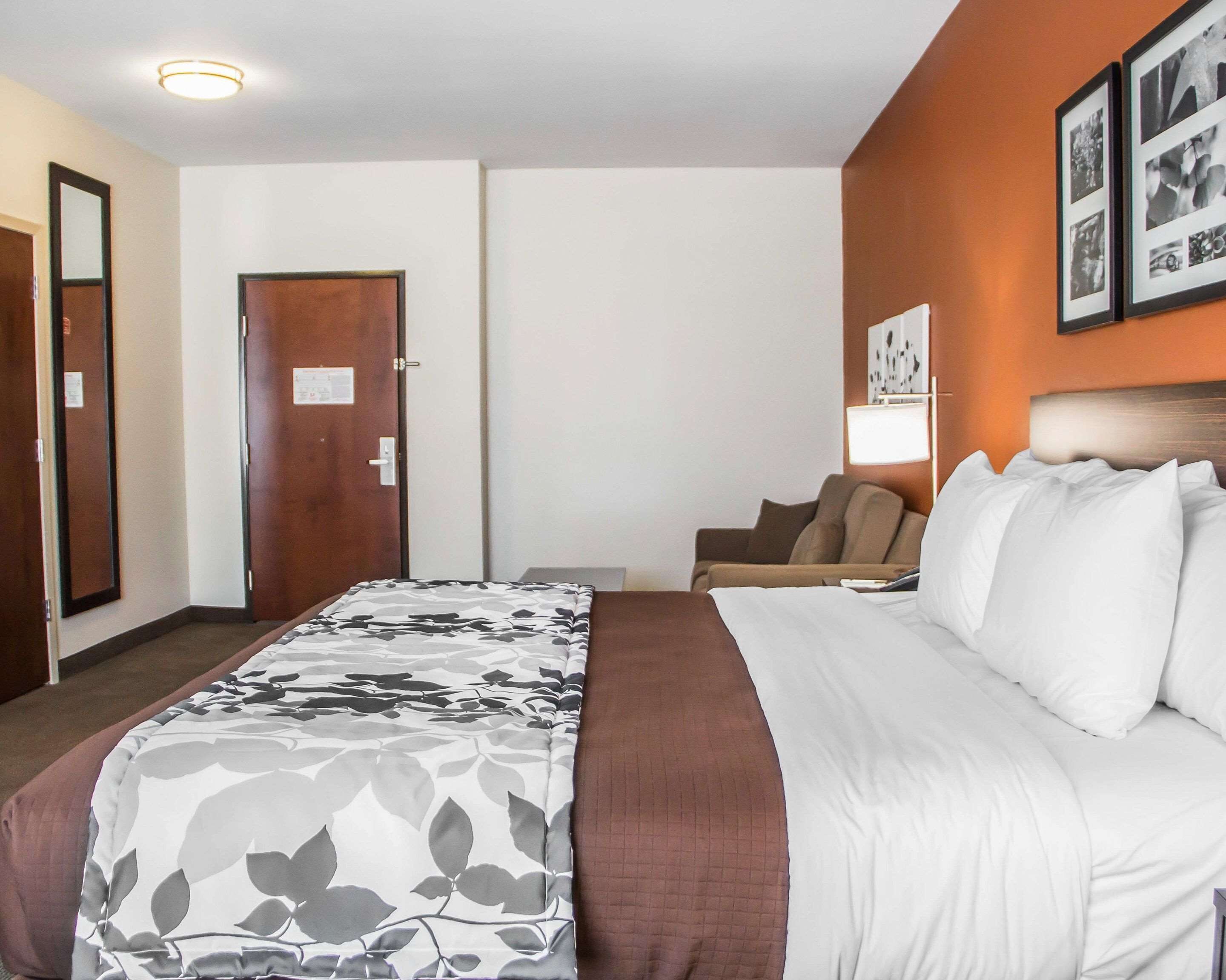 A hotel room with a large bed, a desk, a chair, and a wall-mounted lamp.