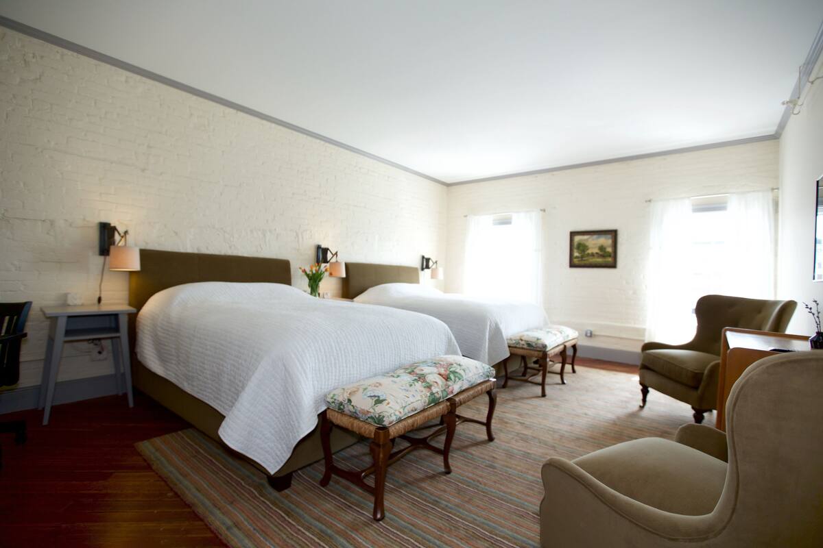 A hotel room with two beds, a desk, a chair, and a small table.