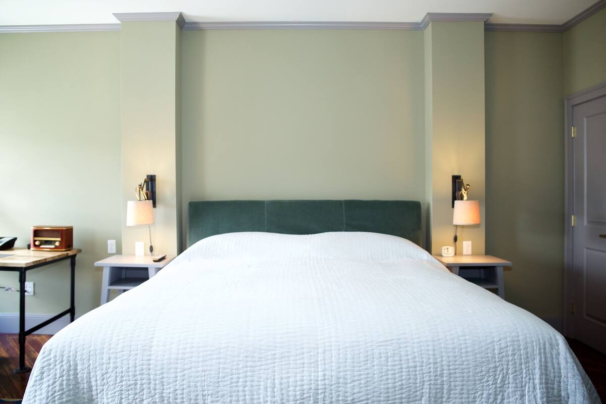 A hotel room with a large bed, two bedside tables with lamps, a small table with a radio, and a door.