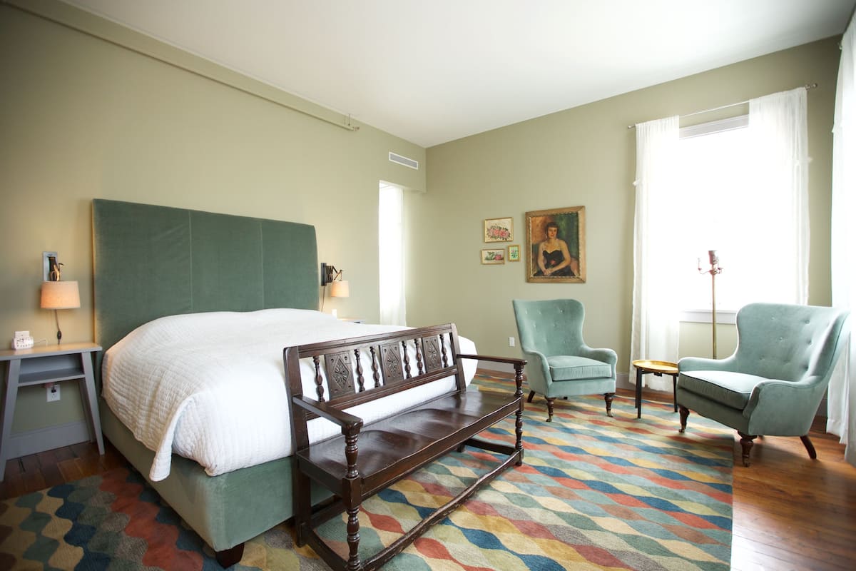 A hotel room with a bed, a bench, a chair, a small table, and a lamp.