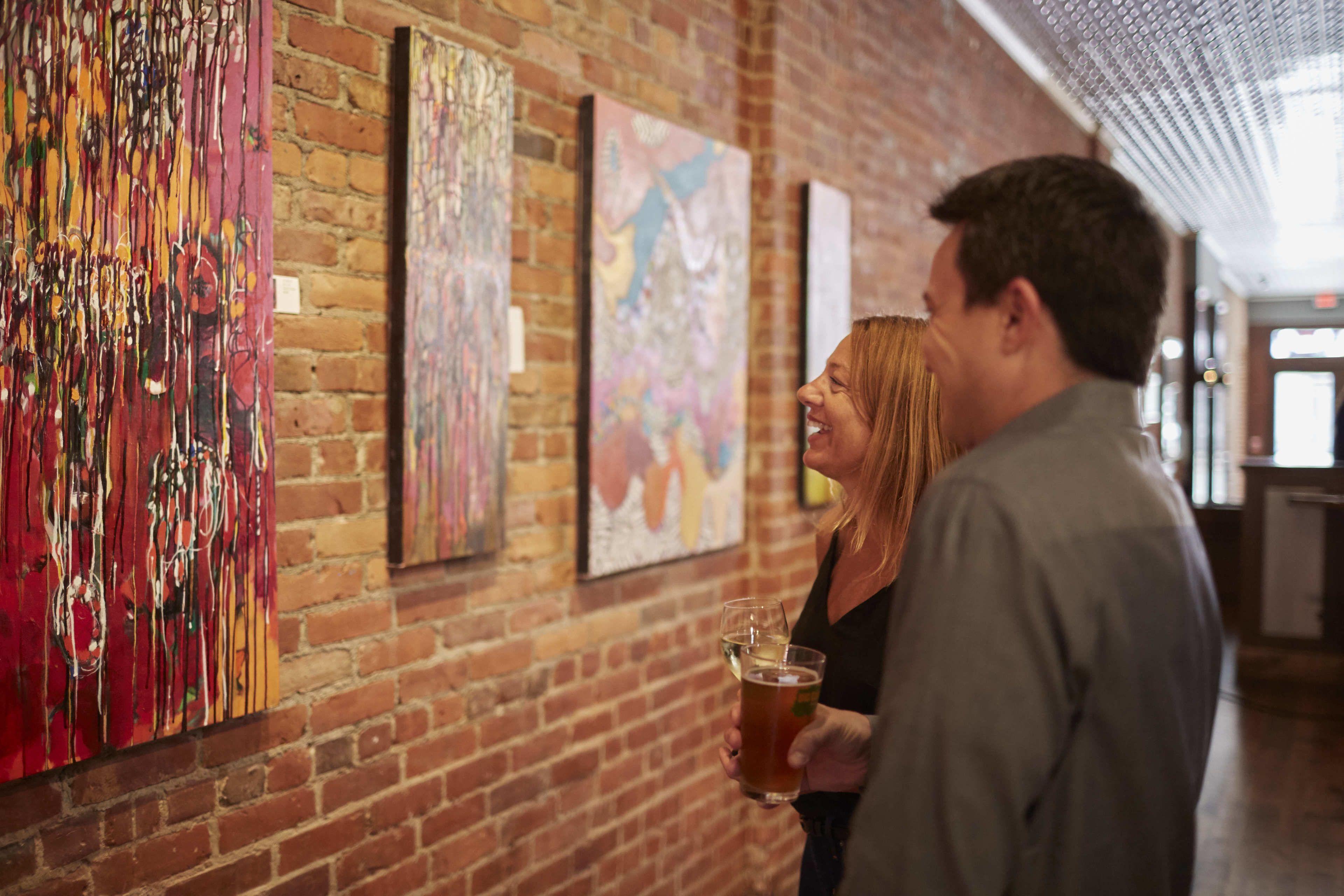 A brick wall with abstract paintings and a person holding a glass.