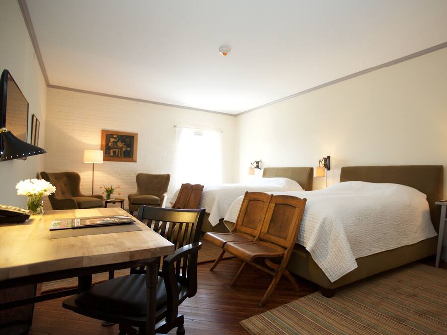A hotel room with a wooden dining table, two wooden chairs, a bed with a white bedspread, a nightstand with a lamp, and a framed picture on the wall.