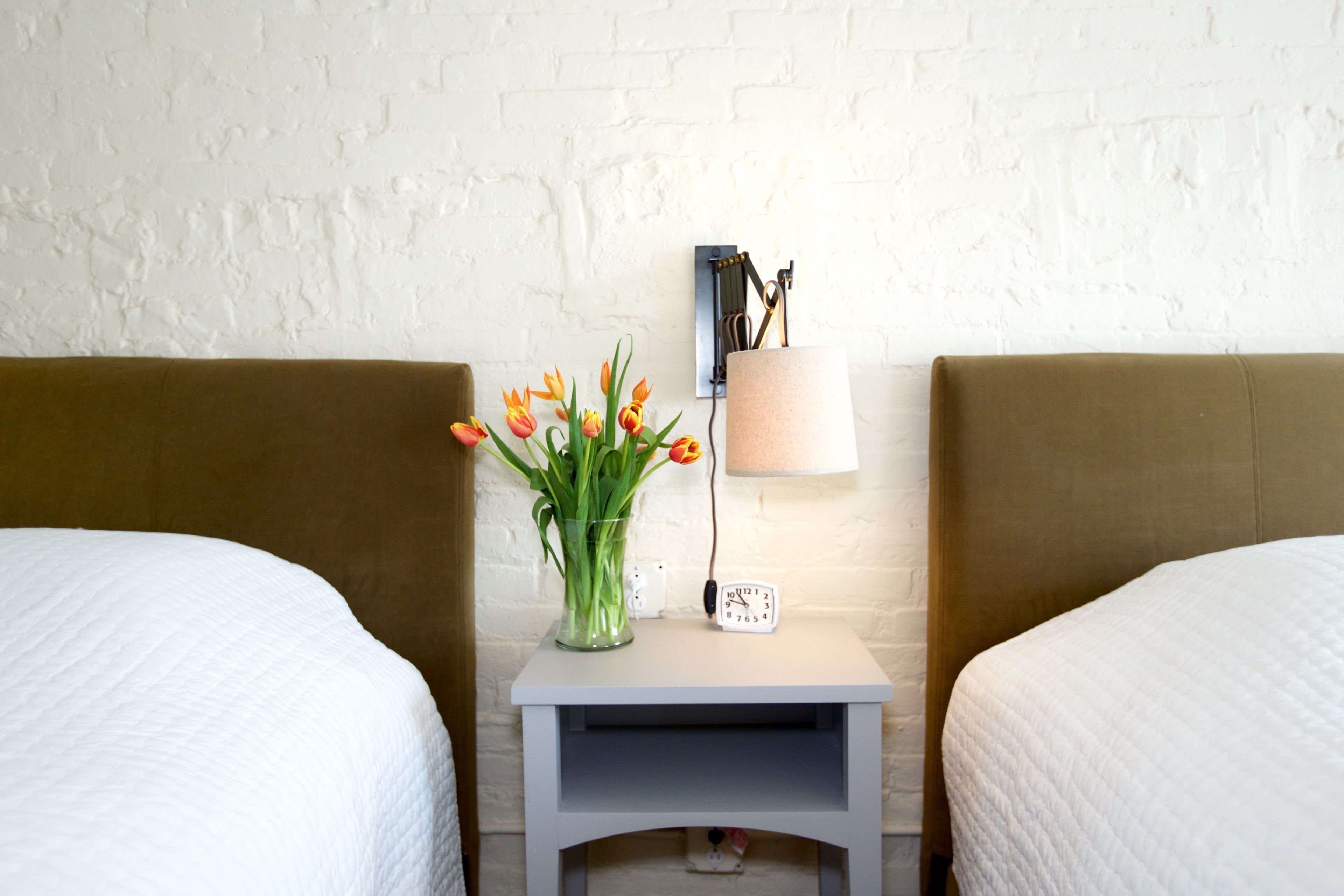 Una habitación de hotel con dos camas, una mesita de noche con una lámpara, un reloj y un florero con flores.