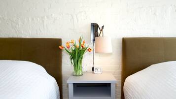 Una habitación de hotel con dos camas, una mesita de noche con una lámpara, un reloj y un florero con flores.