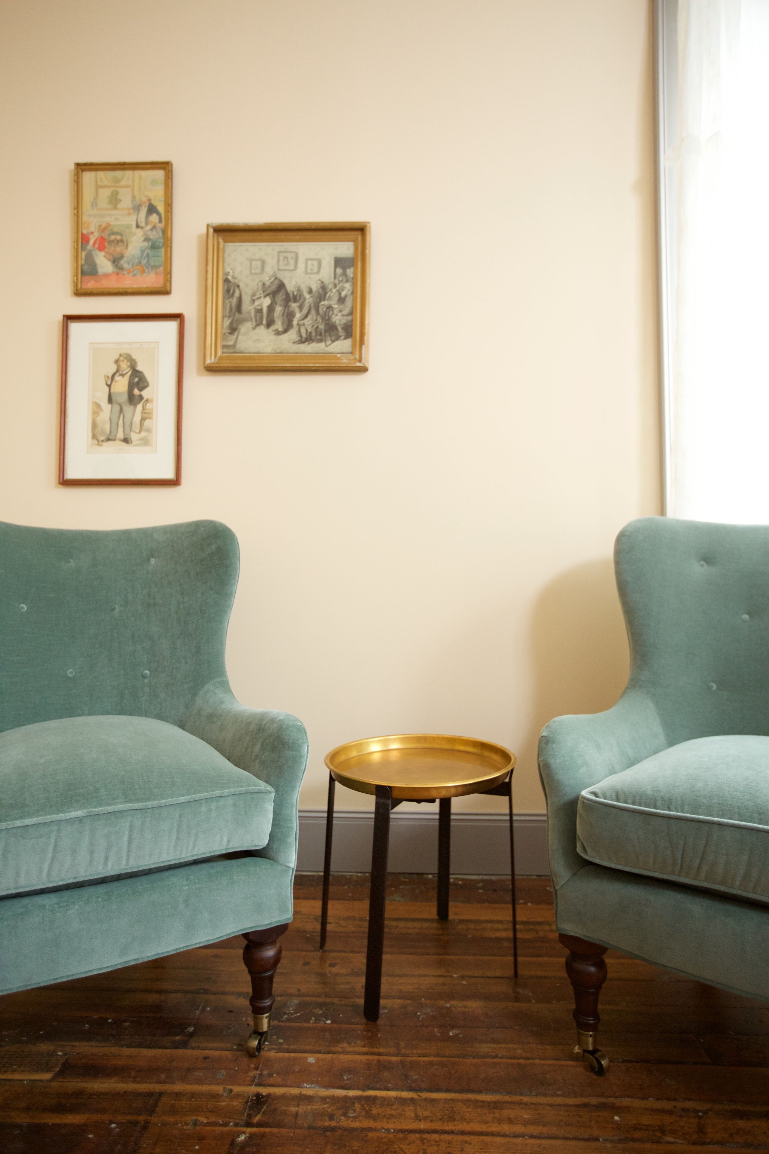 A room with two teal armchairs, a small round table, and three framed artworks on the wall.