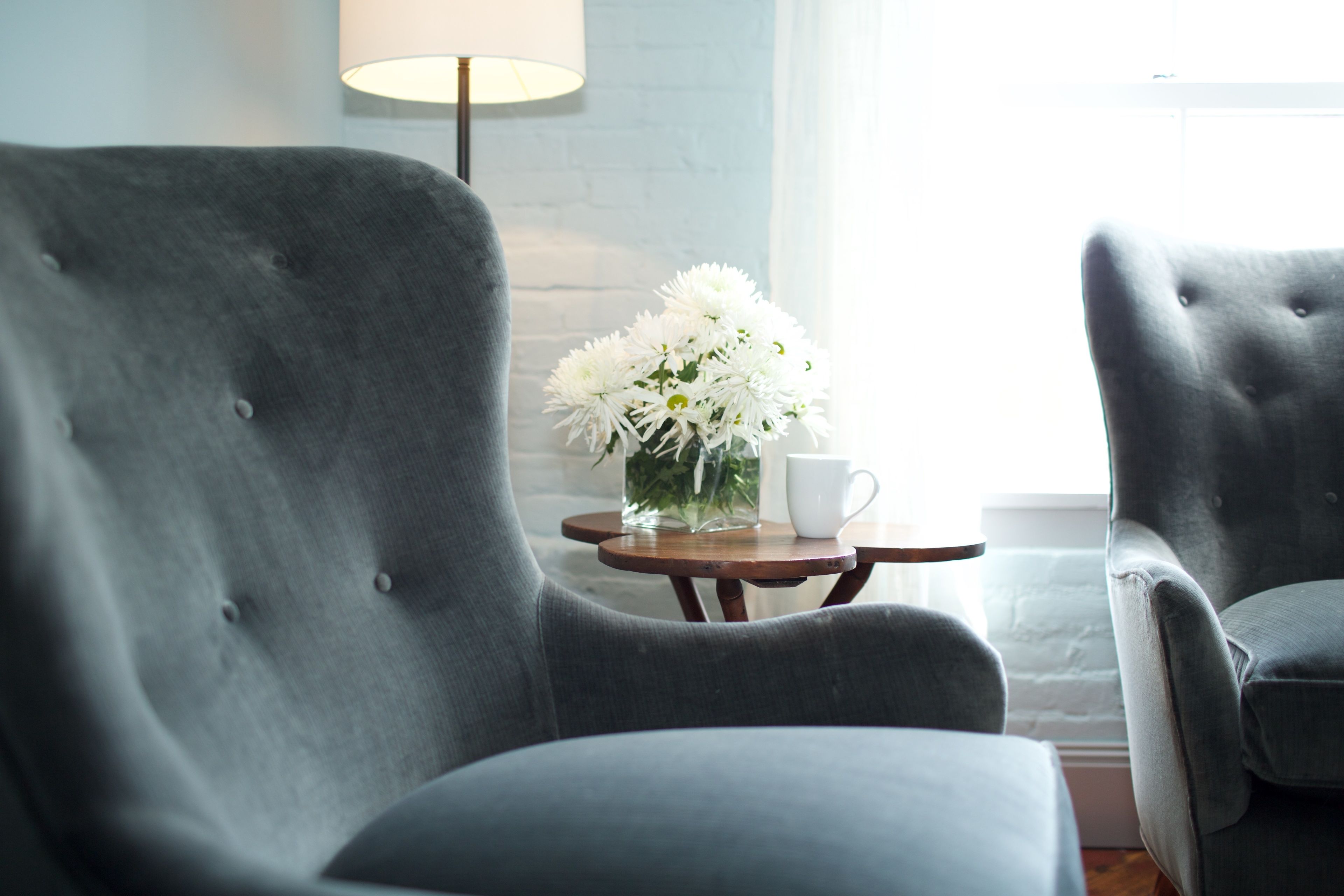 A room with two grey upholstered armchairs, a small wooden table with a white cup and a vase of white flowers, and a lamp in the background.