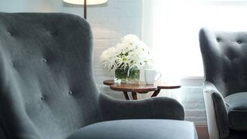 A room with two grey upholstered armchairs, a small wooden table with a white cup and a vase of white flowers, and a lamp in the background.