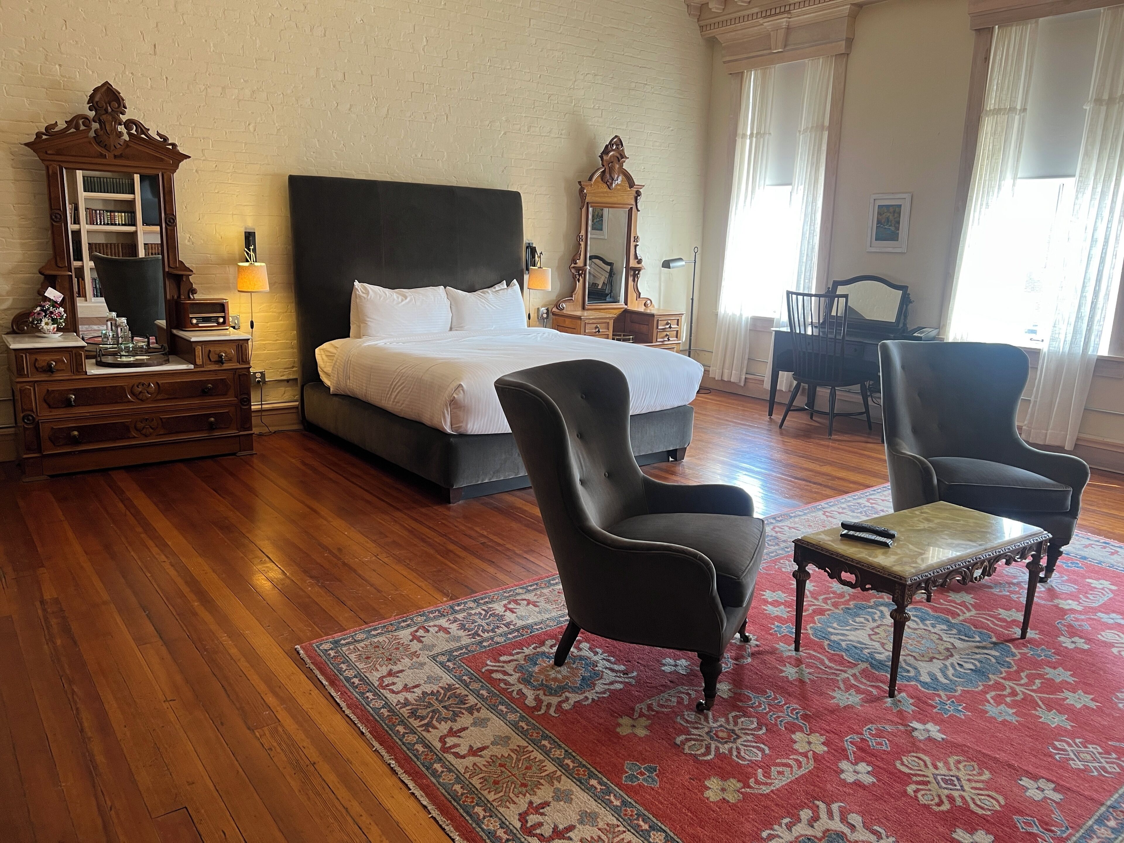 A bedroom with a large bed, two armchairs, a wooden dresser, a mirror, and a piano in the background.