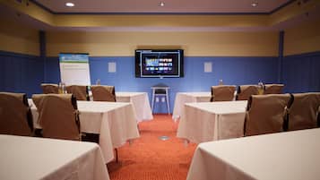 Un salón de conferencias con mesas, sillas, una pantalla de proyector y una pizarra para presentaciones.