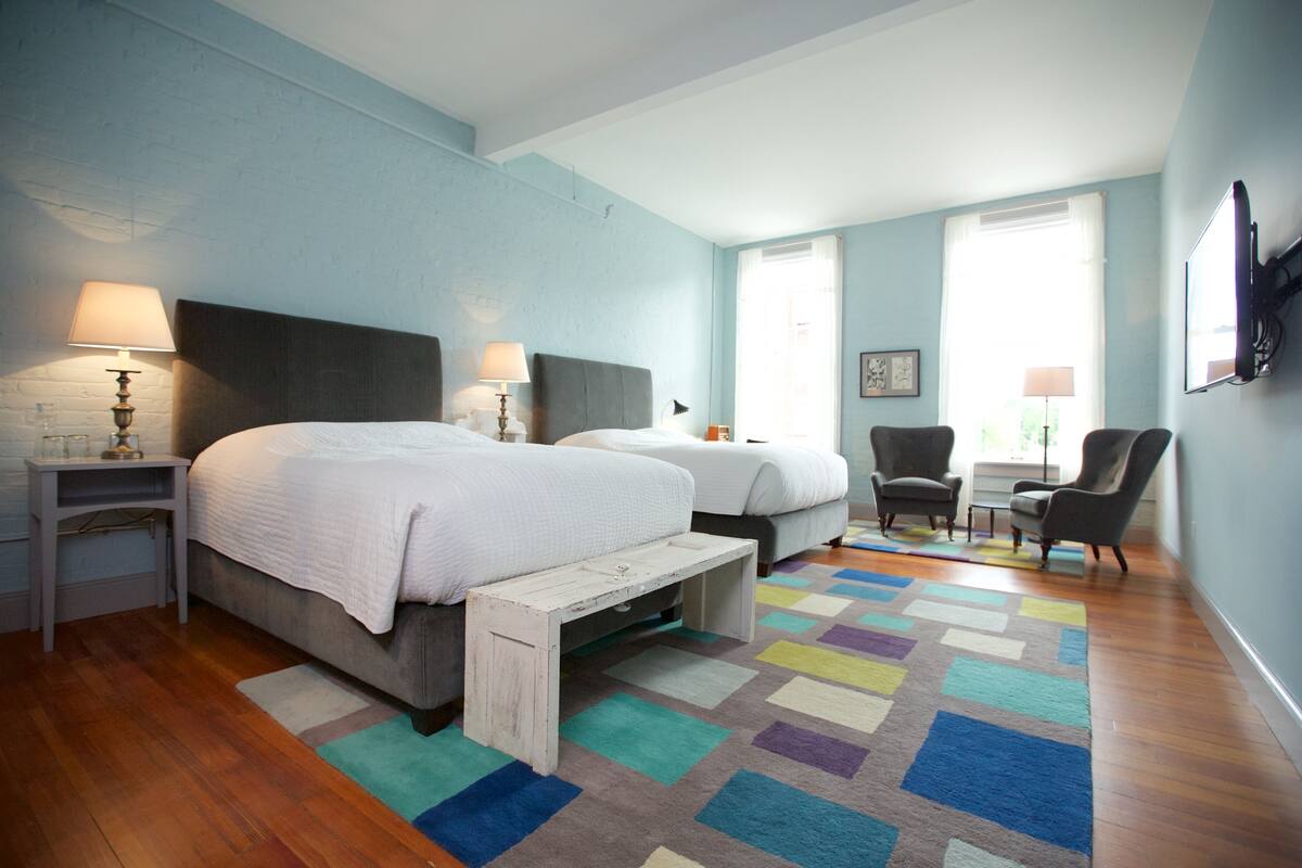 A hotel room with two beds, a small table, two chairs, a colorful rug, and a television on the wall.