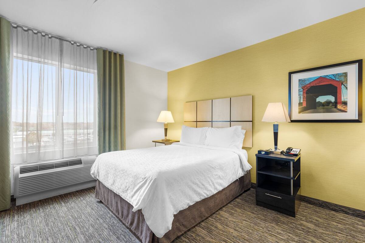 Una habitación de hotel con una cama grande, dos lámparas de noche, un calentador, una mesita de noche con un teléfono y un cuadro en la pared.