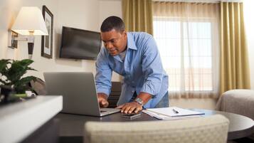 A person using a laptop in a hotel room with a television, a lamp, and a window with curtains.