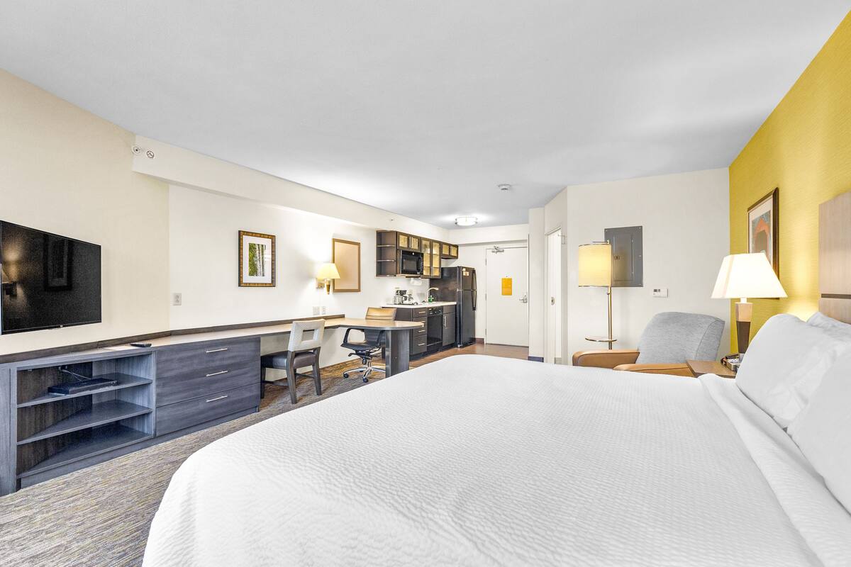 Una habitación de hotel con una cama grande, un escritorio con silla, televisión y kitchenette.